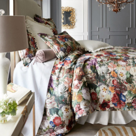 Side shot of a bed with floral bedding, a nightstand with a lamp, plant, and book, with a chandelier and wall mirror in the background