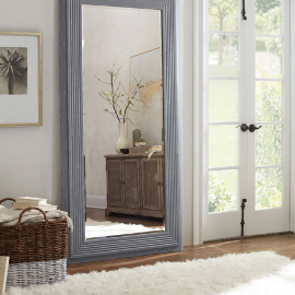 A light blue, wide floor-length mirror in a bedroom with a white, fuzzy rug, next to a framed photo and a basket with laundry in it. In the reflection is a wooden dresser with a white vase and miscellaneous items on top. On the left are French doors leading outside.