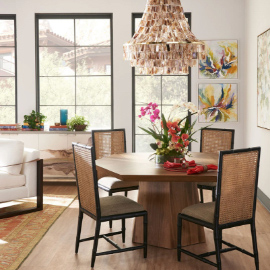 A wooden, hexagon-shaped table surrounded by four rattan chairs in a dining room, with a colorful, floral centerpiece and chandelier. In the background is an armchair, gray/marble chest, decor, and three floor-to-ceiling windows