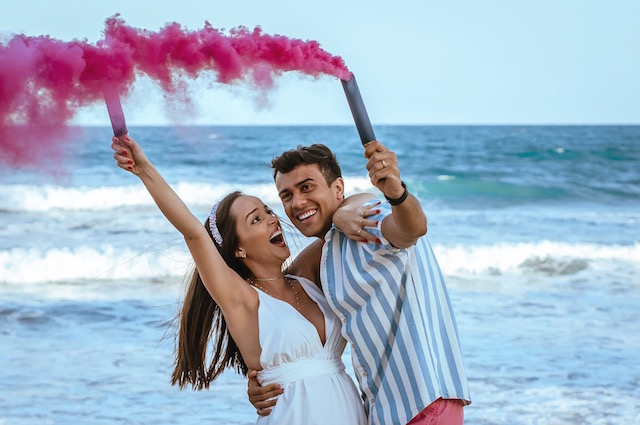 How to Have a Virtual Gender Reveal, a couple standing in front of the ocean on a beach, releasing red smoke out of pipes.