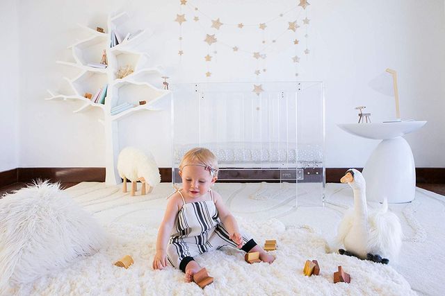 Spring Nursery Trends from Modern Nursery, A baby in black and white striped dress sitting in a nursery with a white rug, white walls, and white, tree shaped shelf