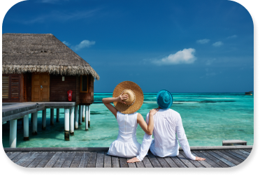 Young couple dressed in white sit on dock looking out onto clear blue ocean with a hut jetting out into the water to the left of couple.