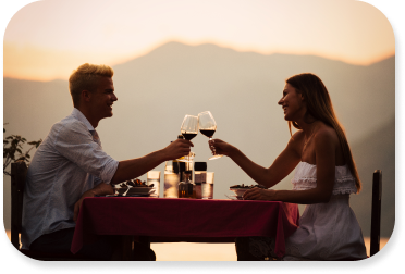 Young couple dressed in white and sitting at table cheers wine glasses as the sun sets.