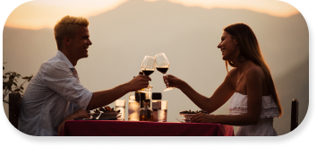 Young couple dressed in white and sitting at table cheers wine glasses as the sun sets.