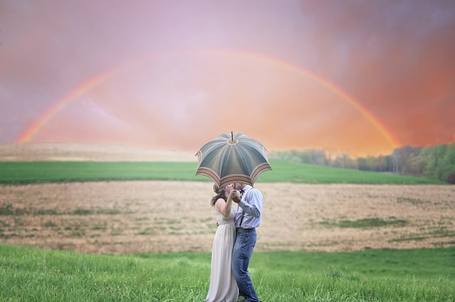Eco-Friendly Wedding Gifts to Celebrate Earth Day, a couple standing in a field, kissing, standing under a parisol. In the sky is a long, beautiful rainbow.