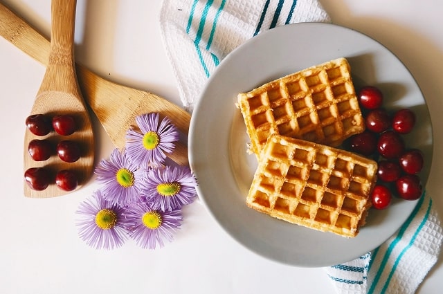 The Best Kitchen Tools to Add to Your Registry, a table populated with a wooden spoon filled with berries, purple flowers, a plate with waffles on it, and cloth napkins.
