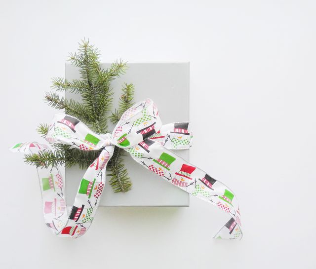 Refinery 29's Guide to Cheap Gifts that Look Anything But, a gray gift box tied with a white ribbon covered with red, green, and black top hats and a small evergreen tree branch.